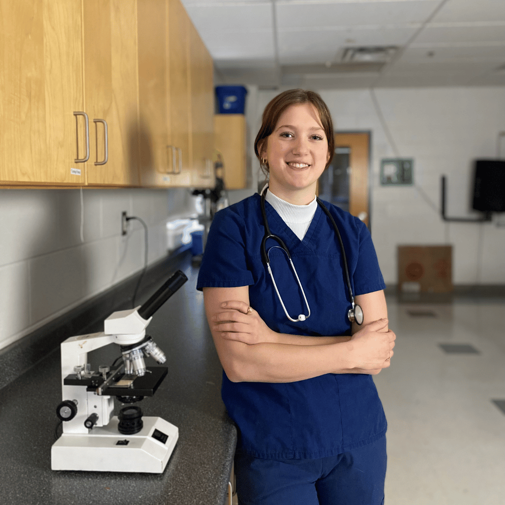 A London Christian High Grade 12 medical cooperative education student posing wearing blue scrubs, a stethoscope, and posing next to a microscope.